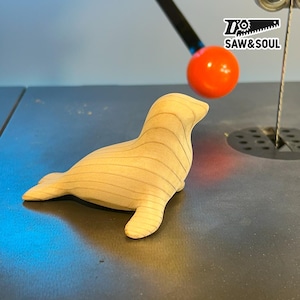 May include: A wooden seal figurine with visible wood grain. The seal is light brown and lying down. The image includes a band saw and an orange ball. The logo "SAW & SOUL" is in the upper right corner.