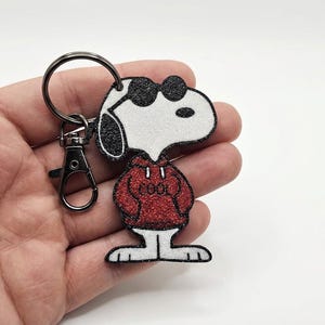 Może przedstawiać: Brelok do kluczy Snoopy z czarnym metalowym karabińczykiem i kółkiem. Postać Snoopy'ego jest biała z czarnymi akcentami, nosi okulary przeciwsłoneczne i czerwoną bluzę z kapturem z napisem "COOL". Zabawny dodatek.
