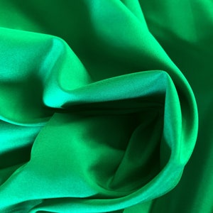 May include: A vibrant emerald green fabric with a smooth, lustrous finish. The material is draped in soft, flowing folds that create deep shadows and bright highlights across the surface, emphasizing the reflective quality of the textile.