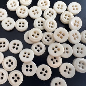 May include: A collection of off-white, round buttons with four holes each. The buttons are scattered on a dark surface, showcasing their simple, classic design. These buttons are suitable for various sewing and craft projects.