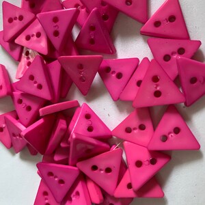Pode incluir: Um monte de botões de plástico rosa em forma de triângulo com dois furos cada. Os botões têm um acabamento brilhante.