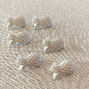 May include: Six white plastic pineapple buttons. The buttons are about 1 inch in diameter and have a raised pineapple design.