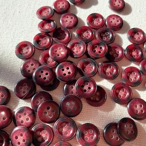 Puede incluir: Una gran colección de botones de color rojo oscuro con un borde negro y cuatro agujeros. Los botones están dispuestos en un patrón aleatorio sobre una superficie blanca.