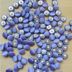 May include: A large group of purple satin buttons with silver backs. The buttons are scattered on a light wood surface.