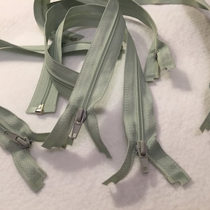 Puede incluir: Cinco cierres de cremallera de color verde claro con tiradores de metal plateado. Las cremalleras están todas abiertas y dispuestas sobre una superficie blanca.