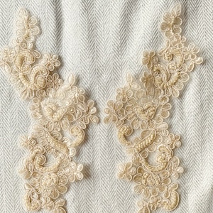 Może przedstawiać: Dwa delikatne, beżowe aplikacje z koronki z kwiatowym wzorem. Aplikacje wykonane są z cienkiej, przezroczystej tkaniny z misternymi detalami i koralikami.