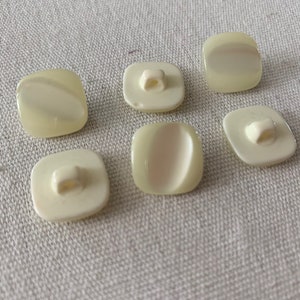 May include: Six white plastic buttons with a square shape and a small hole in the center. The buttons have a slightly raised, oval-shaped design on the front.