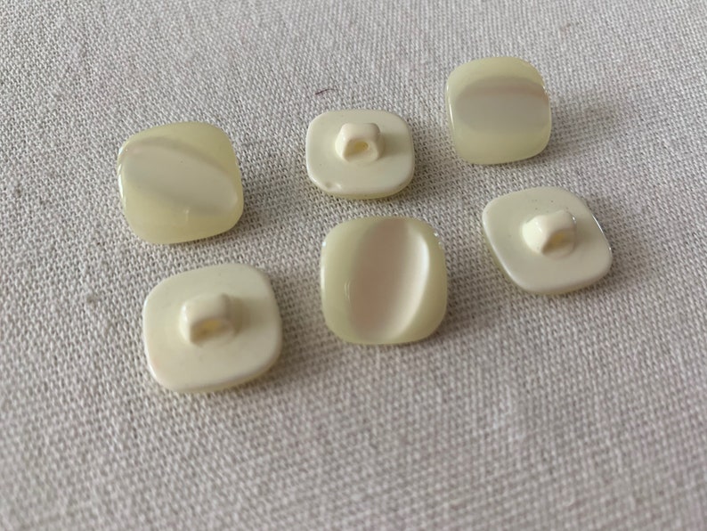 May include: Six white plastic buttons with a square shape and a small hole in the center. The buttons have a slightly raised, oval-shaped design on the front.