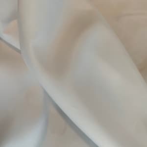 May include: Close-up of a white fabric with soft folds and subtle shadows. The fabric appears smooth and possibly silky, with a slight sheen. The folds create a sense of depth and texture.