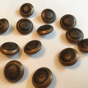 Puede incluir: Un conjunto de botones de color bronce con un diseño de sol. Los botones tienen un diseño circular en relieve en el centro y una forma ligeramente abovedada. Los botones están dispersos sobre una superficie blanca.