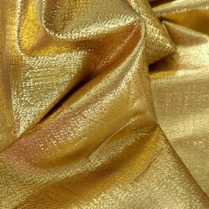 Puede incluir: Primer plano de una tela dorada brillante con una superficie texturizada. La tela est&aacute; doblada, creando sombras y reflejos que realzan su brillo met&aacute;lico. El material parece estar tejido, con un patr&oacute;n sutil visible.