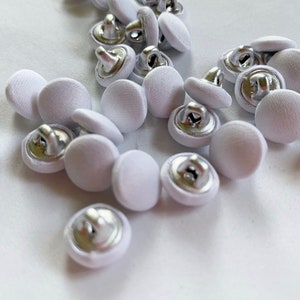 May include: A collection of white fabric covered buttons with silver metal shanks. The buttons are round and have a smooth surface.