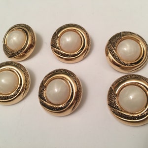 May include: Six gold-toned buttons with a white pearl-like center. Each button has a decorative swirl design around the center.
