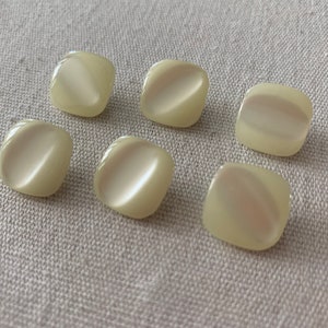 May include: Seven off-white plastic buttons with a rounded square shape. The buttons have a pearlescent finish.