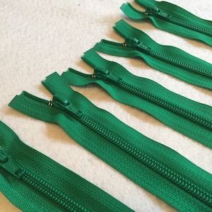 Può includere: Cinque chiusure a zip verdi con cursori in metallo dorato. Le cerniere sono disposte in fila su una superficie bianca.