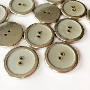 May include: A group of 12 silver-toned buttons with a white center. The buttons have two holes each.