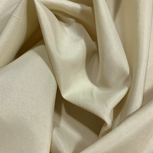 May include: A close-up of a smooth, ivory-colored fabric with a subtle sheen. The fabric is draped and folded, showcasing its soft texture.