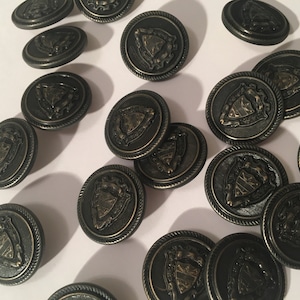 May include: A collection of 17 black metal buttons with a gold-colored design. Each button features a raised crest with a shield and other decorative elements.