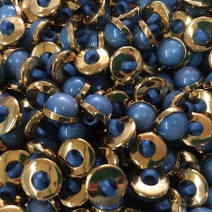 Puede incluir: Un montón de botones azules y dorados. Los botones son redondos y tienen un acabado dorado brillante. Son perfectos para coser o proyectos de manualidades.
