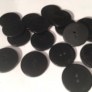 May include: A collection of black, round buttons with two holes each. The buttons are made of a smooth, glossy material and are scattered across a white surface. These buttons are suitable for sewing and craft projects.