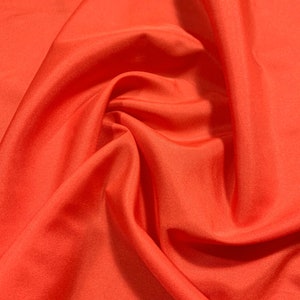 May include: A close-up of a bright orange fabric with a smooth, silky texture. The fabric is draped and folded, creating soft, flowing lines.