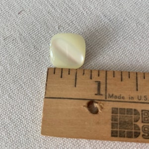May include: A white, square-shaped button with a swirl pattern. The button is about 1 inch in diameter and is shown next to a ruler.