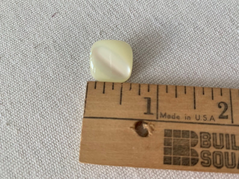May include: A white, square-shaped button with a swirl pattern. The button is about 1 inch in diameter and is shown next to a ruler.