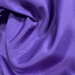 May include: A close-up of a smooth, purple fabric with a slight sheen. The fabric is folded and draped, creating soft folds and curves.