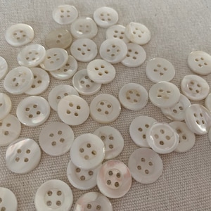 May include: A collection of white mother of pearl buttons with four holes each. The buttons are arranged in a random pattern on a white fabric background.