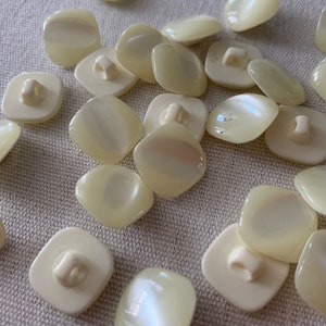 May include: A collection of off-white, square-shaped buttons with a pearly sheen. The buttons have two holes and a shank on the back.
