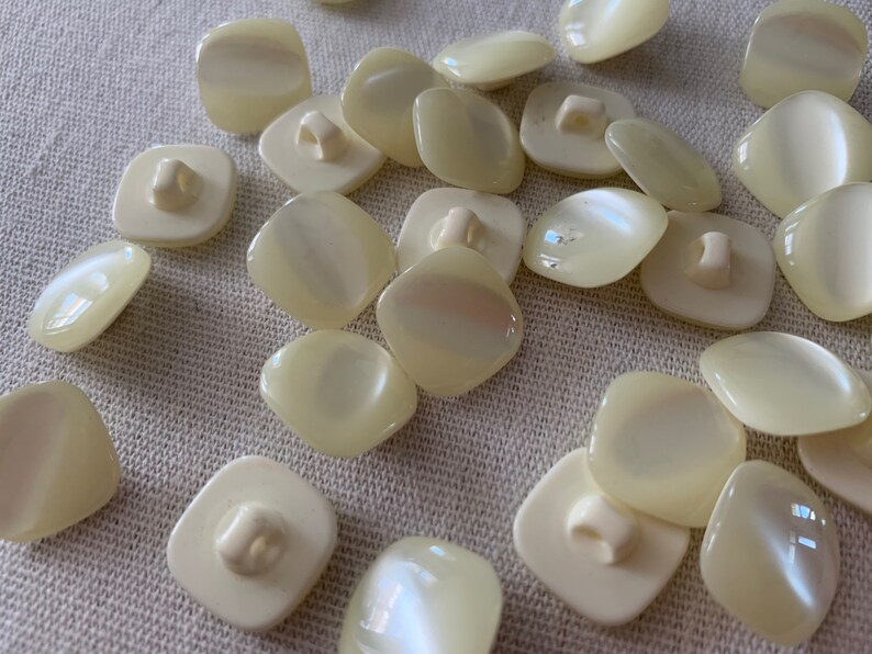 May include: A collection of off-white, square-shaped buttons with a pearly sheen. The buttons have two holes and a shank on the back.