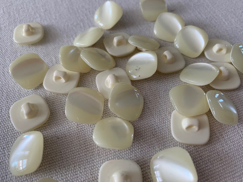 May include: A collection of off-white, square-shaped buttons with a pearly sheen. The buttons have a two-hole design.