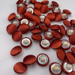 Puede incluir: Una colección de botones cubiertos de tela roja con un centro plateado y blanco. Los botones están esparcidos sobre una superficie blanca.