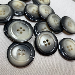 May include: A collection of large, round buttons with a four-hole design. The buttons have a two-tone color scheme, with a dark gray outer ring and a light cream-colored center. The buttons are made of a glossy material.