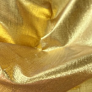 Puede incluir: Primer plano de una tela dorada brillante con una superficie texturizada. El material est&aacute; doblado, creando sombras y reflejos que enfatizan su brillo. La tela parece ligera y fluida.