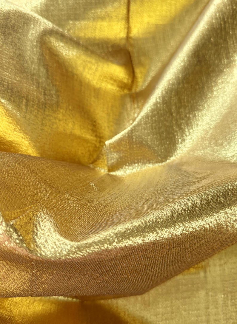 Puede incluir: Primer plano de una tela dorada brillante con una superficie texturizada. El material est&aacute; doblado, creando sombras y reflejos que enfatizan su brillo. La tela parece ligera y fluida.