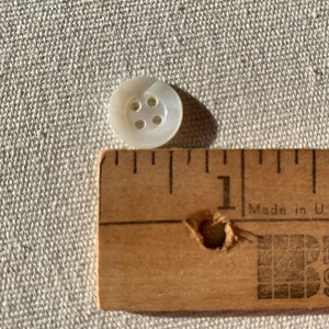 May include: A white mother of pearl button with four holes. The button is about 1/2 inch in diameter and is next to a ruler.