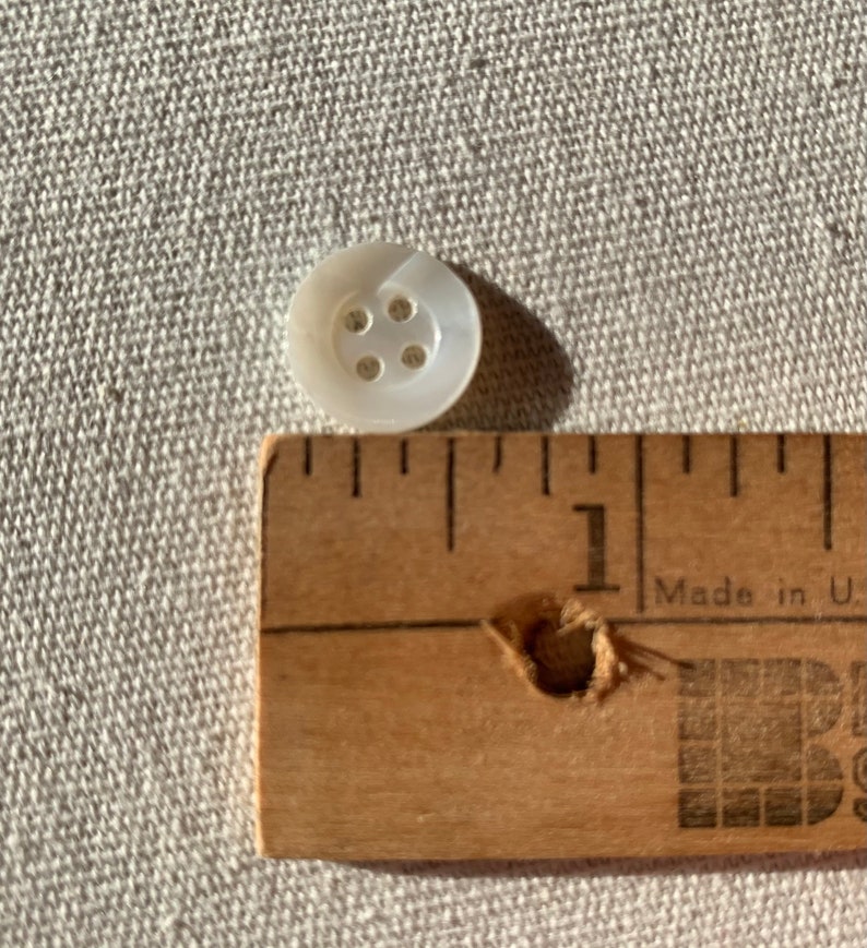 May include: A white mother of pearl button with four holes. The button is about 1/2 inch in diameter and is next to a ruler.