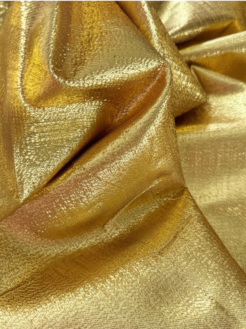 Puede incluir: Primer plano de una tela dorada brillante con una superficie texturizada. La tela est&aacute; doblada, creando sombras y reflejos que realzan su brillo met&aacute;lico. El material parece estar tejido, con un patr&oacute;n sutil visible.
