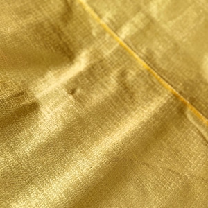 Puede incluir: Primer plano de una tela amarillo dorado con un patr&oacute;n sutil y texturizado. Una fina costura amarilla atraviesa la imagen en diagonal. La tela tiene un ligero brillo, lo que sugiere un material lujoso.