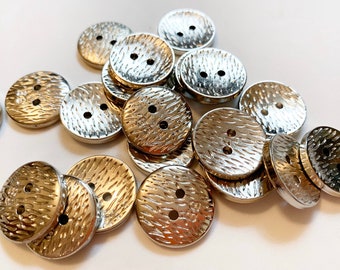 May include: A collection of shiny, silver-coloured buttons with a textured, hammered design. Each button is round with two holes for sewing. The buttons are scattered on a white surface, suitable for sewing projects and crafts.