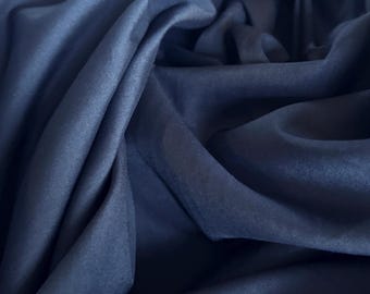 Può includere: Primo piano di un tessuto blu navy con morbide pieghe e una sottile lucentezza. Il materiale appare leggero e fluido, con diverse sfumature di blu che creano profondità e consistenza. Il tessuto è probabilmente utilizzato per abbigliamento o arredamento.