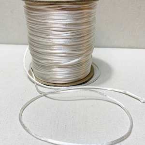 Può includere: Un rocchetto di cordoncino o filo bianco, avvolto attorno a un rocchetto di cartone. Il cordoncino è liscio e lucido, e il rocchetto è marrone.