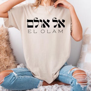 Könnte beinhalten: Beiges T-Shirt mit dem hebräischen Text und der englischen Übersetzung "EL OLAM" in Schwarz gedruckt. Das Shirt hat eine neutrale Farbe und eine lockere Passform.