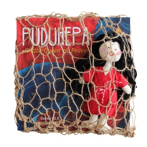 Pu&ograve; includere: Una bambola di stoffa rossa e bianca con capelli neri &egrave; avvolta in una rete marrone. La bambola &egrave; seduta sopra un libro intitolato "PUDUNHEPA White Queen of Peace" di Demet Kilic.