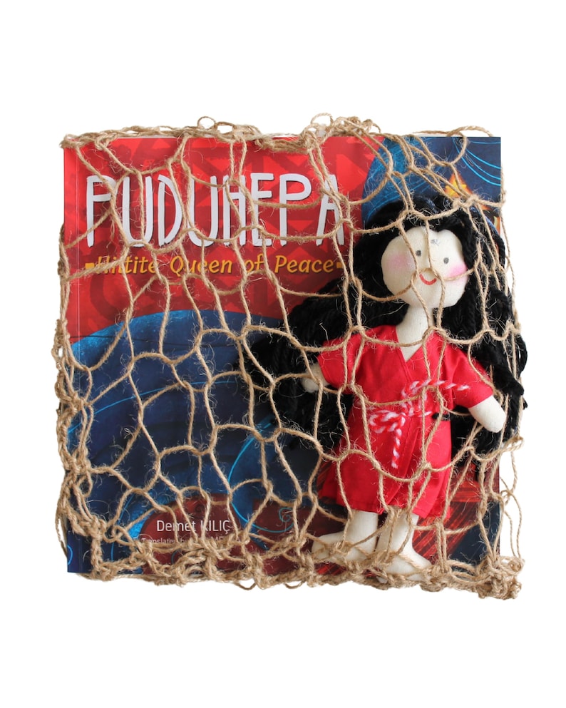 Pu&ograve; includere: Una bambola di stoffa rossa e bianca con capelli neri &egrave; avvolta in una rete marrone. La bambola &egrave; seduta sopra un libro intitolato "PUDUNHEPA White Queen of Peace" di Demet Kilic.