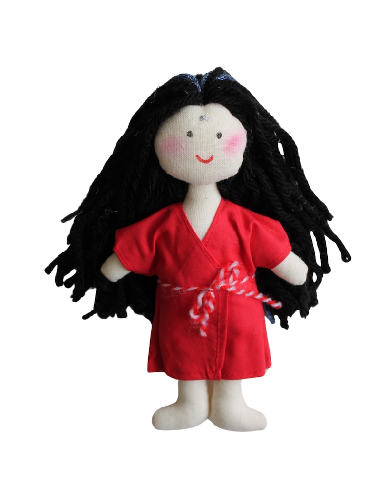 Pu&ograve; includere: Una bambola fatta a mano con lunghi capelli di lana nera, che indossa un kimono rosso con una cintura bianca e rossa. La bambola ha un viso bianco con guance rosa e un semplice sorriso.
