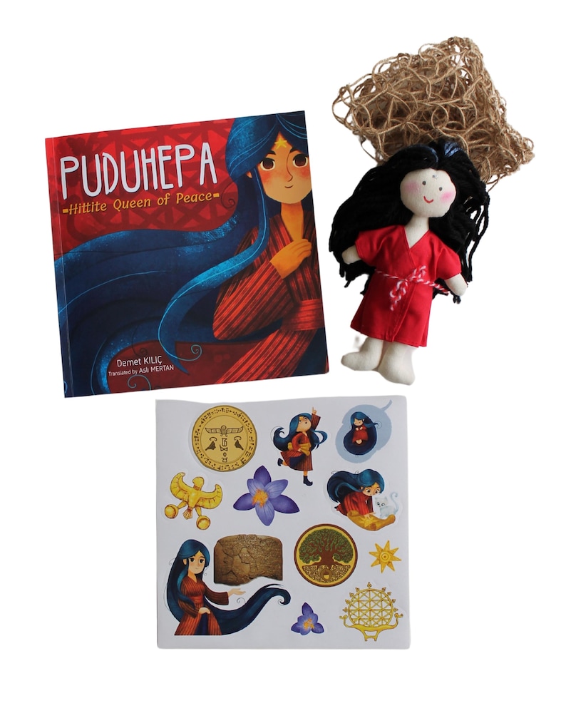 Pu&ograve; includere: Un libro per bambini intitolato "PUDUHЕРА - Hittite Queen of Peace" di Demet Kilic, illustrato da Asli Mertan, una bambola di stoffa rossa con capelli neri e un foglio di adesivi con illustrazioni di una donna con lunghi capelli neri, un albero, un sole e altri simboli.