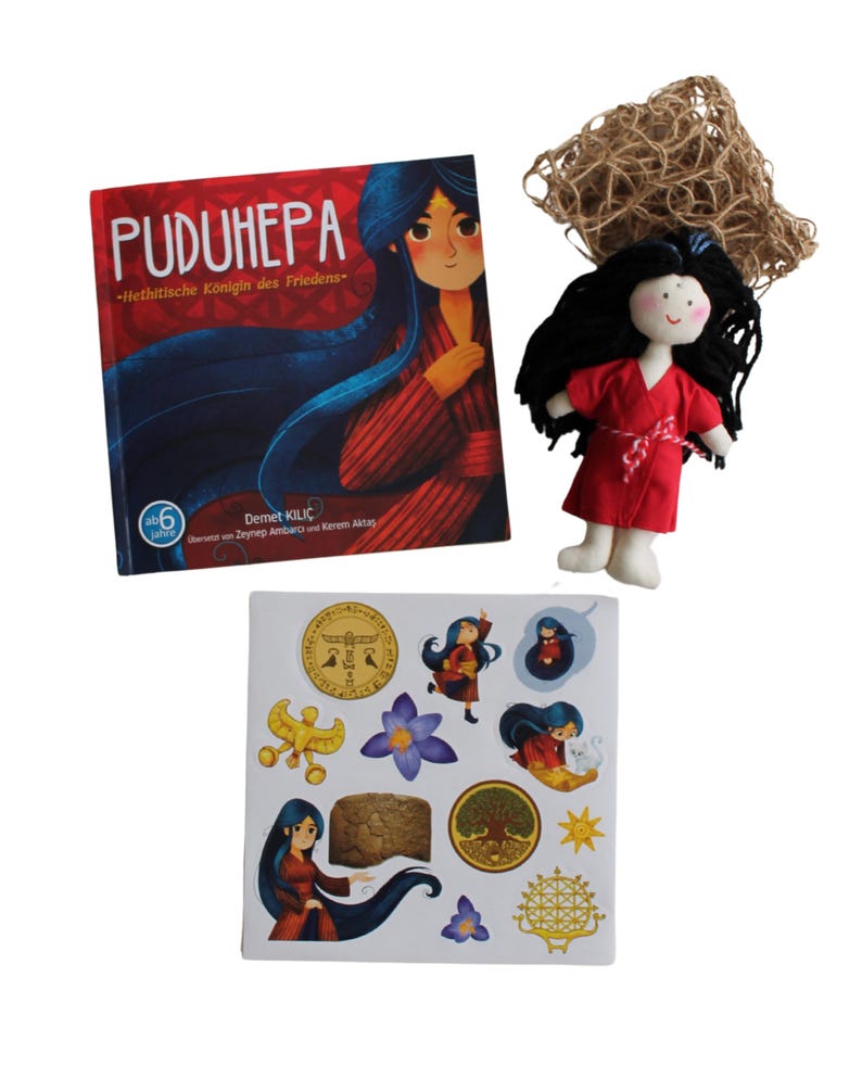 May include: A book titled "Puduhepa - Hittite Queen of Peace" with a colorful illustration of a woman on the cover. A small red fabric doll with black hair and a book of stickers with illustrations of the woman and other symbols.