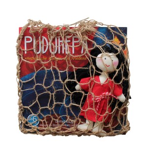 May include: A red and white doll with black hair is inside a brown burlap net bag. The bag is over a book with the title "PUDURHEPA" and the text "Hethitische Konigin des Friedens".